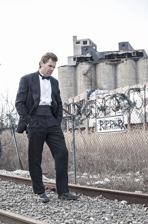 Depressed Caucasian man in formal black tuxedo walks along railroad tracks in urban industrial zoneの写真素材