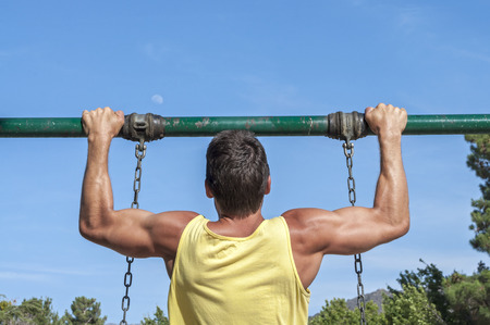 Back view of muscular man in yellow tank top performing pull up exercise on playground swing...