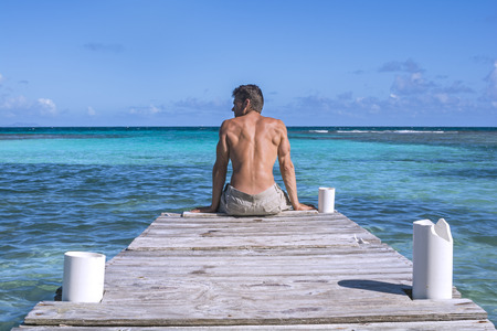 Muscular shirtless Caucasian man sits on edge of dock over beautiful blue green tropical waters of Playa Larga on the Caribbean island of Isla Culebra in Puerto Ricoの写真素材