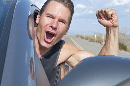 Closeup of handsome free Caucasian man driving car with head out window down desert road as he laughs with extreme excitement and waves armの写真素材