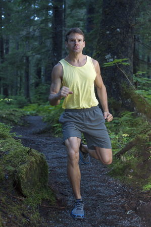 Muscular Caucasian man running in short and tank top on trail through dense forestの写真素材