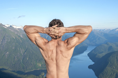 Back of shirtless muscular Caucasian man stretching arms with fingers locked behind head as he enjoys the magnificent scenic view over body of water surrounded by snow capped mountains near Sitka, Alaskaの写真素材