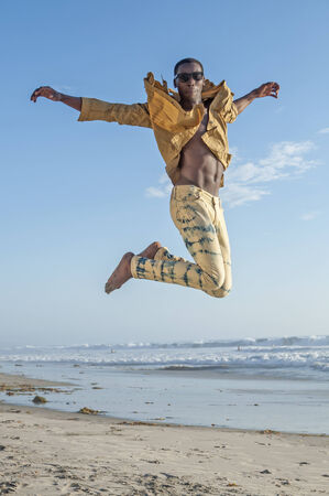 Fashionable sexy African American man leaps high in air on beach as he shows off stylish clothesの写真素材