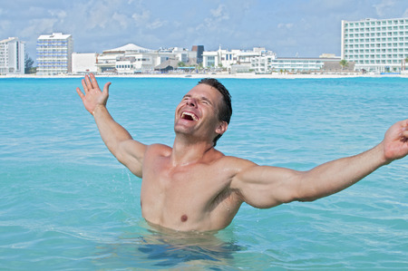 Young fit and shirtless Caucasian man celebrates with arms outstretched in shallow tropical waters of beach resortの写真素材