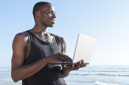 Handsome lean young African American man smiles as he observes activity on beach while connected online with small laptop computer in sunny outdoorsの写真素材