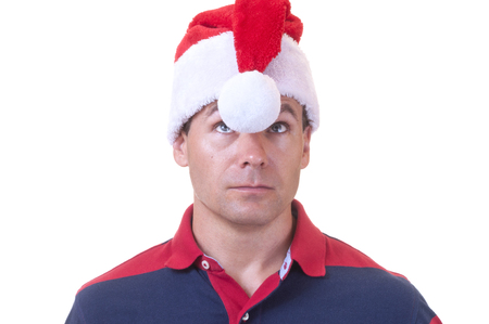 Caucasian man with confused expression in casual polo shirt looks up at white ball hanging between his eyes from red Santa hat on white backgroundの写真素材