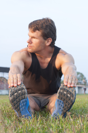 Focus on shoes as muscular Caucasian man sits in grass reaching for toes to do hamstring stretch on grass sport fieldの写真素材
