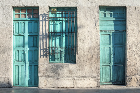 Two aqua green painted wooden doors and window of rustic exterior of old colonial style building illuminated by morning light in Merida, Yucatan, Mexicoの写真素材