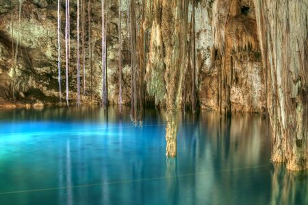 Beautiful clear blue water of Xkenken cenote in Dzitnup, Mexico illuminated from above with tree roots and stalactites hanging from ceiling of cavernの写真素材