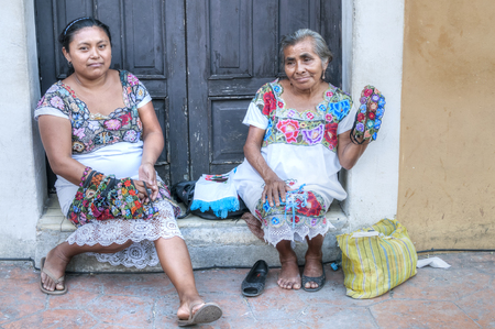 VALLADOLID, MEXICO - JANUARY 20, 2015: Two women in traditional dress sit in front of old rustic building along sidewalk offering textile handicrafts for sale to touristsのeditorial素材