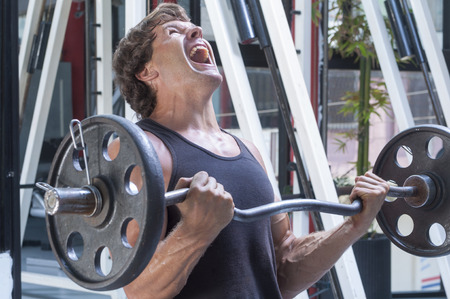 Muscular Caucasian man in tank top painfully struggles to complete repetition of arm curls in gymの写真素材