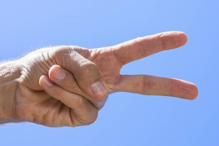 Closeup low angle of male hand showing peace sign with blue sky background on sunny day in natural lightの写真素材