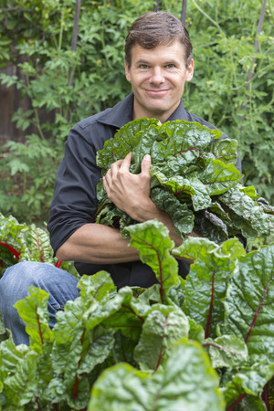 Handsome Caucasian man with big smile as he carries harvest of fresh picked chard leaves in backyard gardenの写真素材