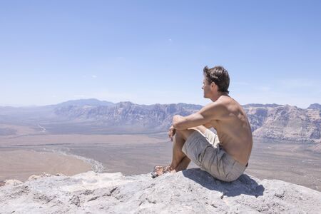 Wide angle of shirtless muscular Caucasian man in shorts and sandals sitting on rocky summit of mountain overlooking expanse of desert under sunny clear sky in Red Rock Canyon, Nevadaの写真素材