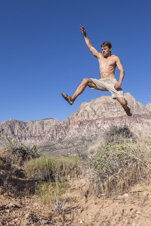 Muscular shirtless Caucasian man wearing shorts and sandals runs and jumps high in air over bushes in harsh desert landscape under clear sunny blue sky in Red Rock Canyon, Las Vegasの写真素材