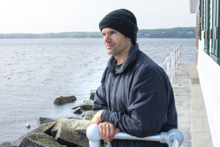 Handsome Caucasian man dressed in jacket and black cap leans against railing of lighthouse at end of breakwater in Rockland Maine as he gazes peacefully into the distant oceanの写真素材