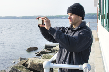 Caucasian man dressed in black jacket and beanie takes cell phone photo of scenic view of bay at the lighthouse at end of breakwater in Rockland Maineの写真素材