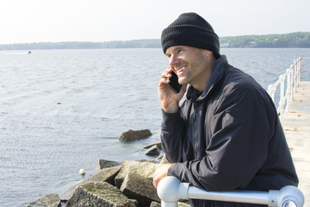 Handsome Caucasian man smiles while talking on cell phone as he leans against railing at end of breakwater of bay in Rockland Maineの写真素材
