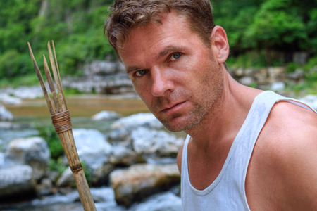 Closeup portrait of handsomely rugged Caucasian man with primitive bamboo spear in natural setting of jungle river environmentの写真素材