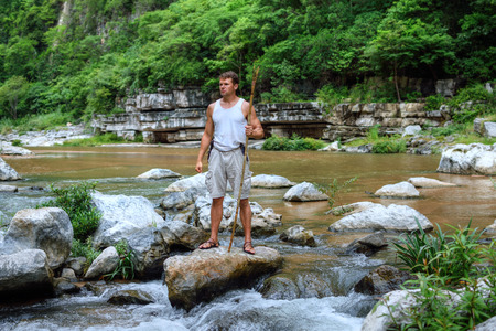 Muscular adventurous Caucasian man in tank top and shorts holding spear while standing triumphantly on boulder in middle of river in tropical jungle in Chiapas, Mexicoの写真素材