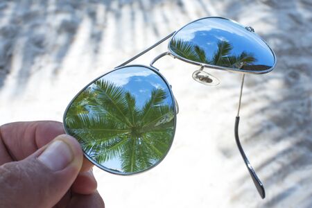 Closeup on lens of reflective sunglasses over sandy beach reveals reflections of tall coconut palms in tropical paradiseの写真素材
