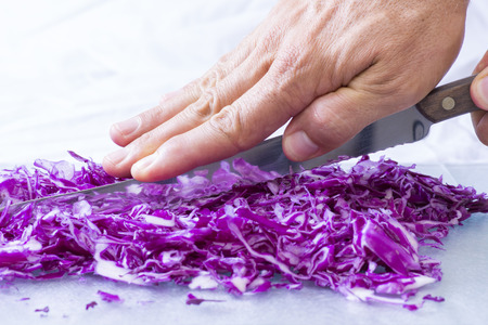 Closeup of hand pressing spine of serrated knife cutting small pieces of red cabbage in brightly lit kitchenの写真素材