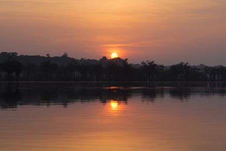 Beautiful vivid orange sky as sun rises over jungle landscape along bank of Urubu River in Amazonas, Brazil and reflects on surface of waterの写真素材