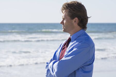 Frustrated and stressed out Caucasian office worker in blue shirt and red tie stands outdoors at beach in warm sunshine and thinksの写真素材