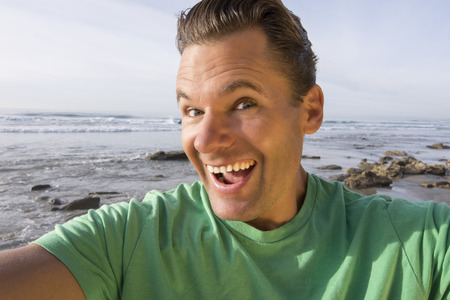 Closeup selfie photo of handsome funny Caucasian man with big silly smile wearing green shirt at beatiful beachの写真素材