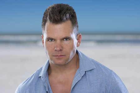 Closeup portrait of handsome Caucasian man wearing blue shirt with serious bold expression of determination on blurred beach backgroundの写真素材