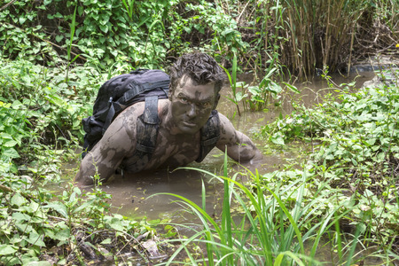 Adventurous man covered in mud wears backpack while hiking through deep muddy water and green...