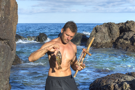 Lean shirtless adventure man holds fresh caught parrotfish and primitive fishing rod on rocky Caribbean shore on Isla Culebraの写真素材