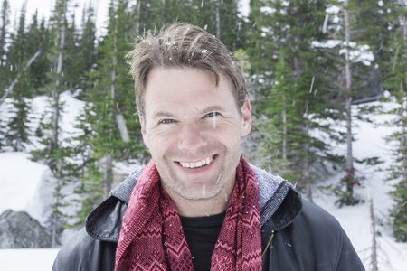 Portrait of smiling handsome Caucasian man wearing black jacket and red scarf while it snows in beautiful pine forestの写真素材