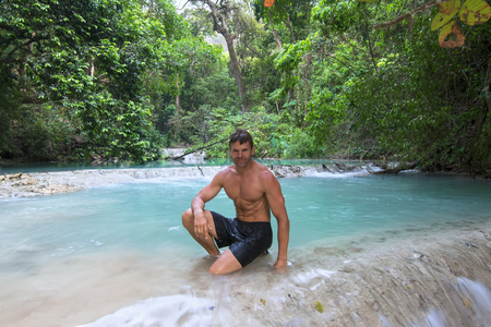 Muscular shirtless Caucasian man kneeling on edge of beautiful turquoise blue pool in lush tropical forest above La Conchuda waterfall in Chiapas, Mexicoの写真素材