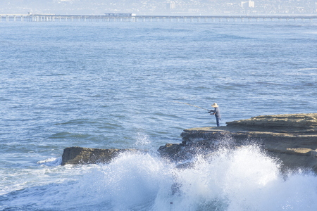 Waves crash against the rocky cliffs of Ocean Beach, California as a surf fisherman fishes from above the splash zone on a summer morningの写真素材