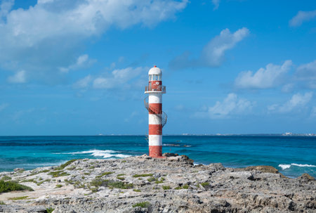 Red and white striped Punta Cancun lighthouse situated on end of rocky point with Isla Mujeres island in backgroundの写真素材