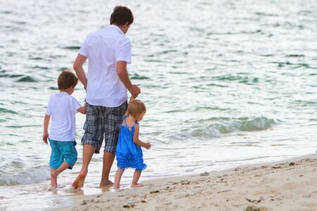 Young father with his two kids walking along tropical white sand beach at evening timeの写真素材