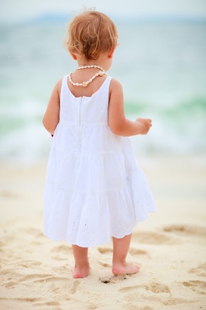 Back view of toddler girl standing at tropical beach facing oceanの写真素材