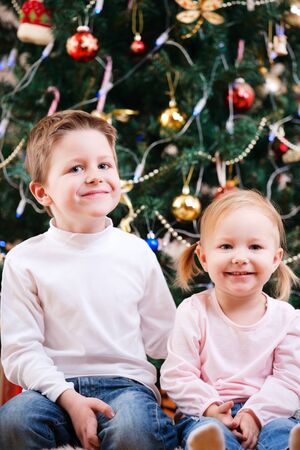 Two small kids sitting near beautifully decorated Christmas treeの写真素材