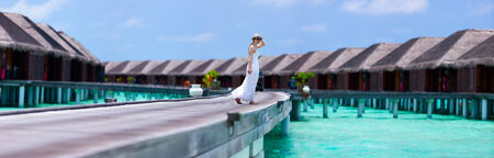 Beautiful girl in a long white dress walking on a wooden jetty at resortの写真素材