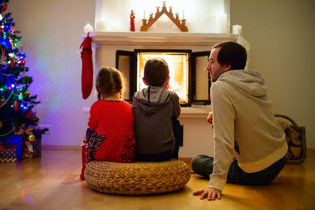 Father and his two little kids sitting by a fireplace in their family home on Christmas eveの写真素材