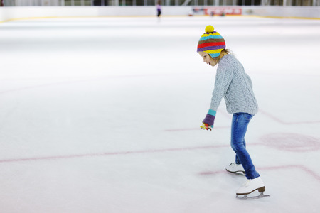 Adorable little girl wearing jeans, warm sweater and colorful hat skating on ice rink