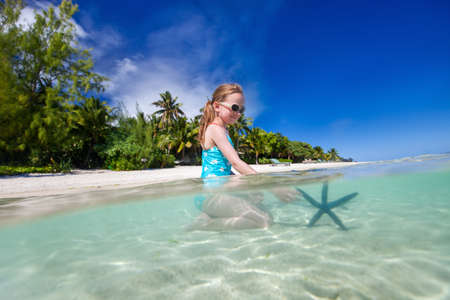 Split underwater photo of a little girl playing with blue starfish on shallow clear ocean waterの写真素材
