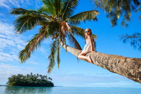 Adorable little girl at tropical beach sitting on palm tree during summer vacationの写真素材