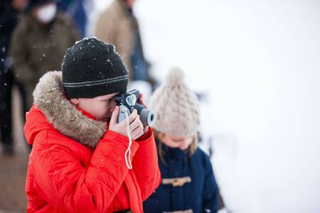 Cute boy in a red parka down jacket outdoors on beautiful winter snow day taking photos with small cameraの写真素材