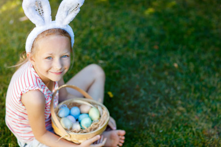 Adorable little girl wearing bunny ears holding a basket with Easter eggs outdoors on spring day