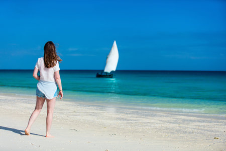 Back view of a young beautiful woman at white sand tropical beach looking at traditional african...