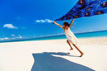Cute little girl having fun running with sarong and enjoying vacation at tropical beach with white sand and turquoise ocean waterの写真素材