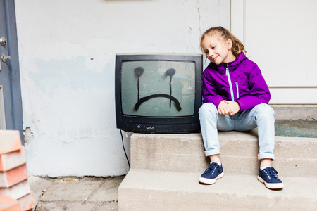 Adorable girl sitting next to vintage TV with sad face drawn on itの写真素材