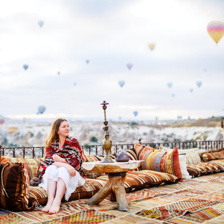 Young woman relaxing on terrace with amazing morning view of hot air balloons flying over Goreme...